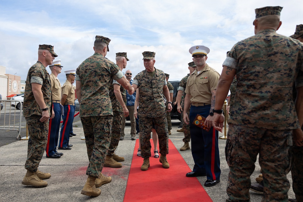 A formal military reception ceremony is taking place on a red carpet outdoors, likely at a U.S. Marine Corps installation. A senior officer in camouflage utilities is being greeted by personnel lined up on both sides of the red carpet. The setting and uniforms indicate this is an official arrival or honors ceremony.