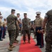A formal military reception ceremony is taking place on a red carpet outdoors, likely at a U.S. Marine Corps installation. A senior officer in camouflage utilities is being greeted by personnel lined up on both sides of the red carpet. The setting and uniforms indicate this is an official arrival or honors ceremony.
