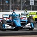 An IndyCar race is in progress on a street circuit, with a blue PPG/Hitachi-sponsored car prominently in the foreground. The driver's name 'McLaughlin' is visible on the cockpit halo. A yellow-green car follows closely behind in second position, with spectators visible behind safety fencing in the background.