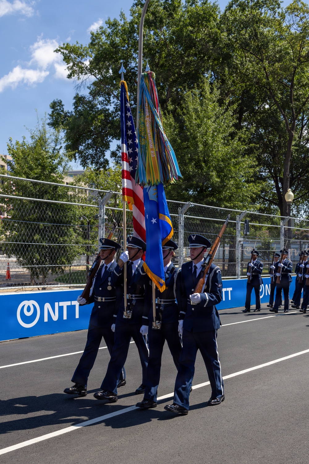 A U.S. Air Force color guard is marching in formation on a paved street, carrying the American flag and the Air Force flag adorned with streamers. The ceremony appears to be taking place at a motorsport event venue, evidenced by the NTT IndyCar Series branding on the barrier walls. Additional uniformed personnel are visible standing at attention in the background.