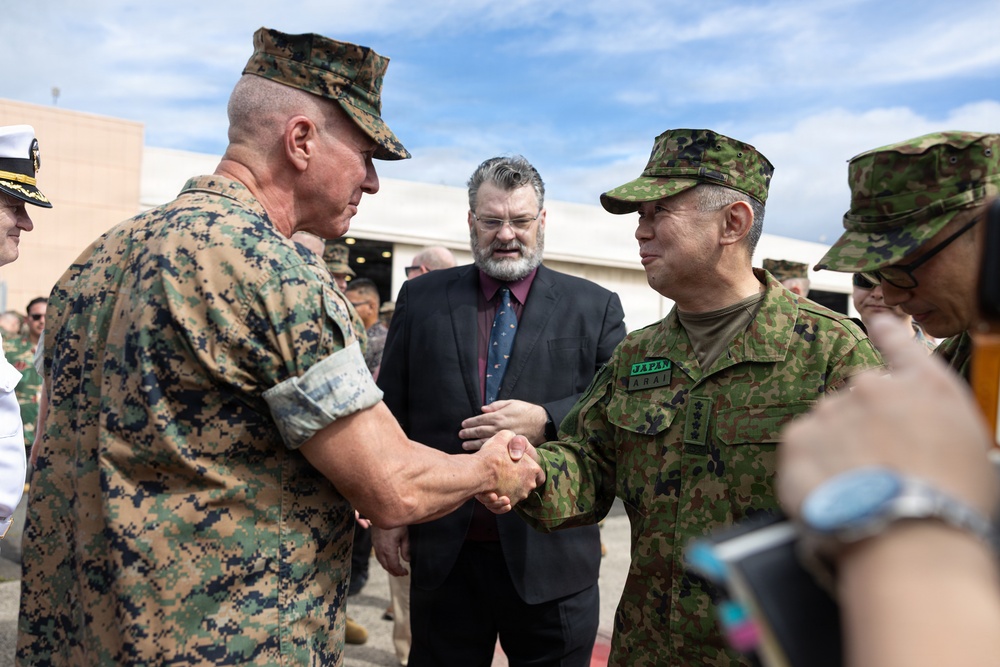 Two military officers are shaking hands in what appears to be an official meeting or greeting ceremony. One officer wears US Marine Corps MARPAT digital camouflage, while the other wears Japanese Ground Self-Defense Force camouflage with a 'JAPAN' and 'ARAI' name tape visible. A civilian in a dark suit with a beard observes the exchange in the background.