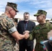 Two military officers are shaking hands in what appears to be an official meeting or greeting ceremony. One officer wears US Marine Corps MARPAT digital camouflage, while the other wears Japanese Ground Self-Defense Force camouflage with a 'JAPAN' and 'ARAI' name tape visible. A civilian in a dark suit with a beard observes the exchange in the background.