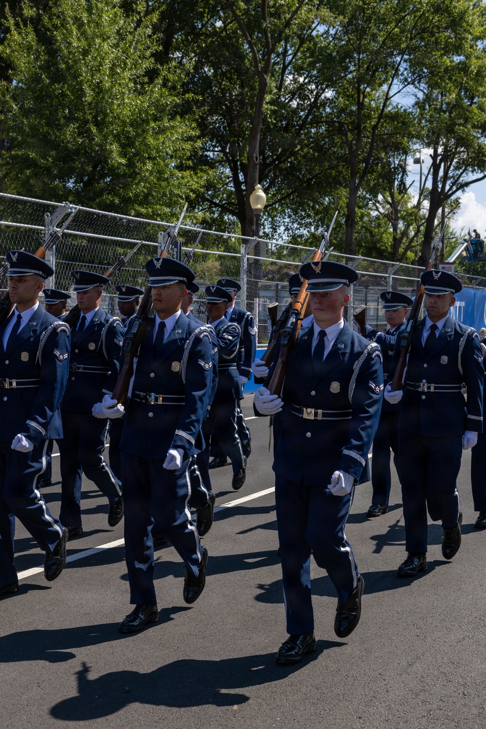 A United States Air Force Honor Guard unit is marching in formation during a parade or ceremonial event. The personnel are dressed in formal dark blue dress uniforms with white gloves and peaked caps, carrying rifles with bayonets at shoulder arms. The event appears to take place on a paved road lined with chain-link security fencing and mature trees in the background.