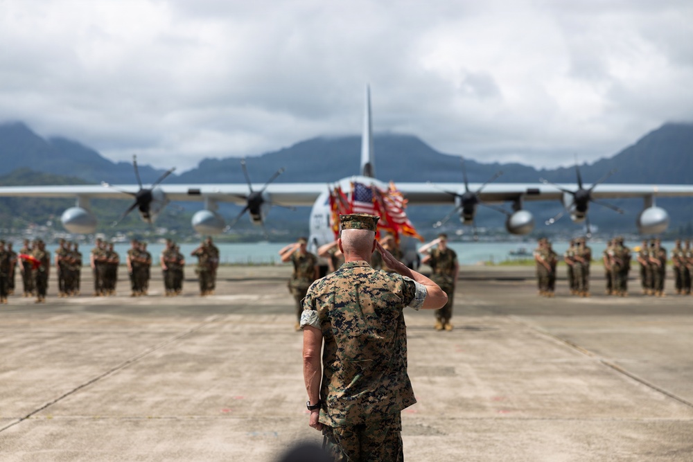 A military ceremony is taking place on an airfield tarmac, with a senior Marine officer saluting a formation of troops. An American flag and what appears to be a unit colors flag are visible among the formation, with a large military transport aircraft (C-130 Hercules) serving as a backdrop. The setting appears to be a coastal airfield with green mountains and water visible in the background, consistent with Hawaii.