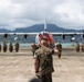 A military ceremony is taking place on an airfield tarmac, with a senior Marine officer saluting a formation of troops. An American flag and what appears to be a unit colors flag are visible among the formation, with a large military transport aircraft (C-130 Hercules) serving as a backdrop. The setting appears to be a coastal airfield with green mountains and water visible in the background, consistent with Hawaii.