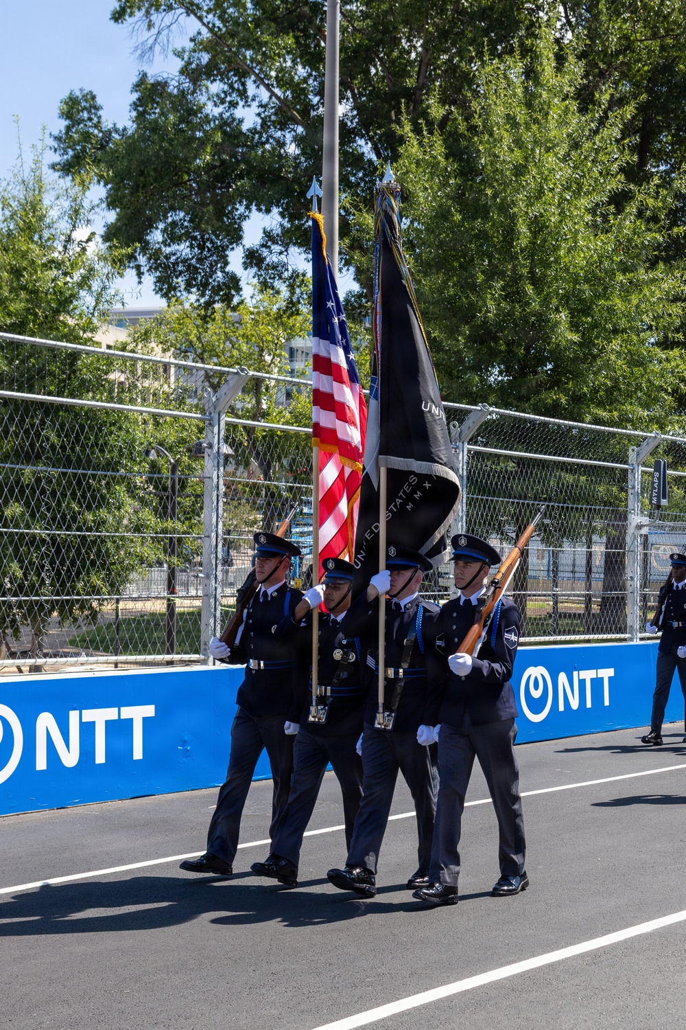 A military color guard unit is marching in formation on what appears to be a racing circuit or street course, carrying the United States flag and a dark ceremonial flag. The personnel are dressed in dark blue dress uniforms with white gloves and peaked caps, with some carrying rifles. Blue barrier walls bearing the 'NTT' sponsor logo line the track, suggesting this is a pre-race ceremony at an IndyCar or similar motorsport event.