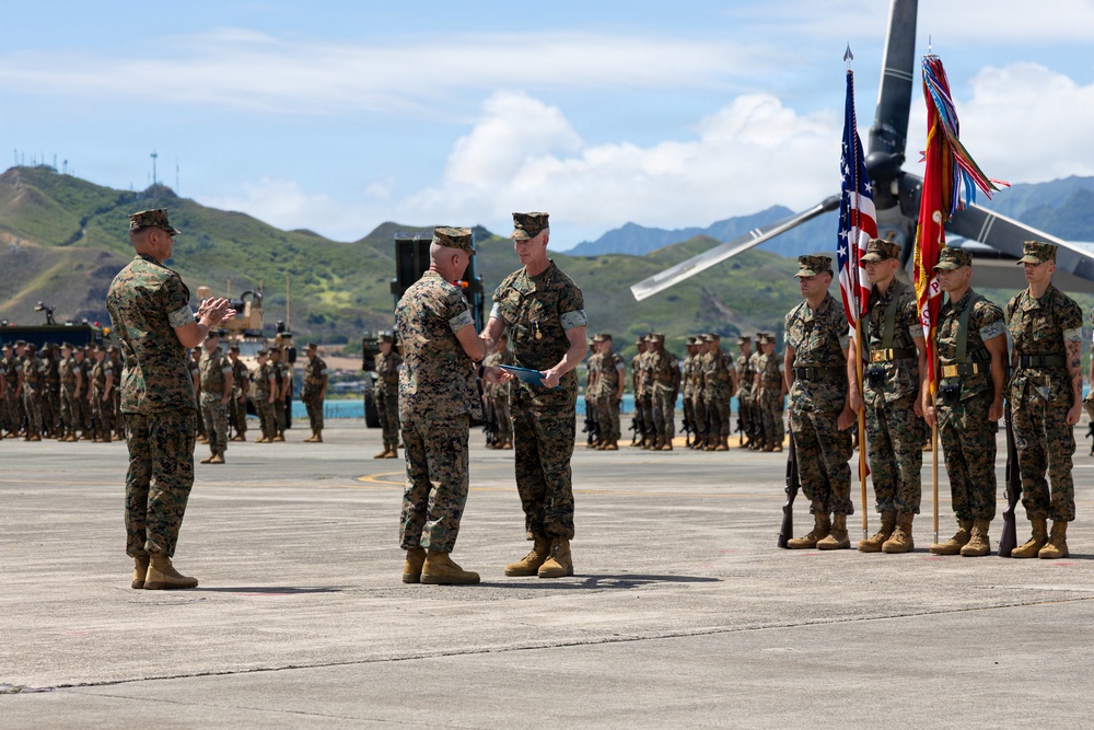 A formal U.S. Marine Corps military ceremony is taking place on a tarmac or airfield, likely a change of command, award presentation, or unit review. Three senior Marines are positioned at the front conducting the ceremony, while a color guard holds flags including the U.S. flag and unit colors, and rows of Marines stand in formation in the background. The setting appears to be in a tropical or coastal location based on the green hills and terrain visible in the background.