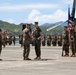 A formal U.S. Marine Corps military ceremony is taking place on a tarmac or airfield, likely a change of command, award presentation, or unit review. Three senior Marines are positioned at the front conducting the ceremony, while a color guard holds flags including the U.S. flag and unit colors, and rows of Marines stand in formation in the background. The setting appears to be in a tropical or coastal location based on the green hills and terrain visible in the background.