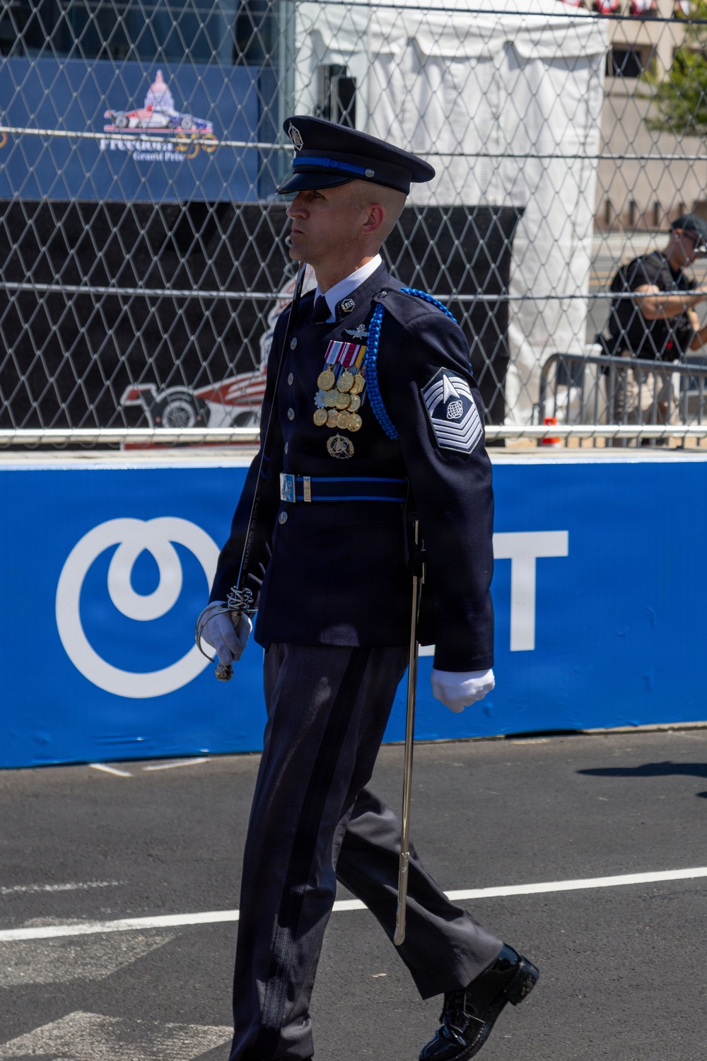 A United States Air Force senior non-commissioned officer in dress blues uniform is marching in a ceremonial parade. The individual displays numerous medals, a blue aiguillette, and a Chief Master Sergeant rank insignia on the sleeve. The background shows signage consistent with the 'Freedom Grand Prix' IndyCar racing event.
