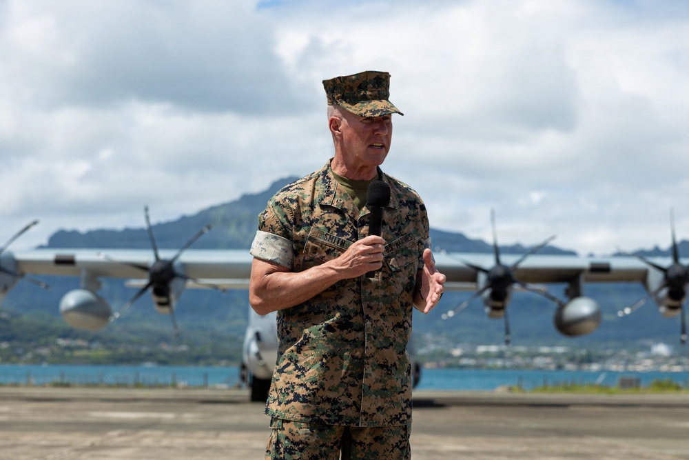 A U.S. Marine Corps officer in MARPAT woodland camouflage uniform is speaking into a microphone on an airfield tarmac. Behind him are military turboprop aircraft, likely C-130 Hercules transports, with a coastal landscape and mountainous terrain visible in the background. The setting appears to be a tropical military air station, consistent with Hawaii.