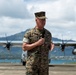 A U.S. Marine Corps officer in MARPAT woodland camouflage uniform is speaking into a microphone on an airfield tarmac. Behind him are military turboprop aircraft, likely C-130 Hercules transports, with a coastal landscape and mountainous terrain visible in the background. The setting appears to be a tropical military air station, consistent with Hawaii.