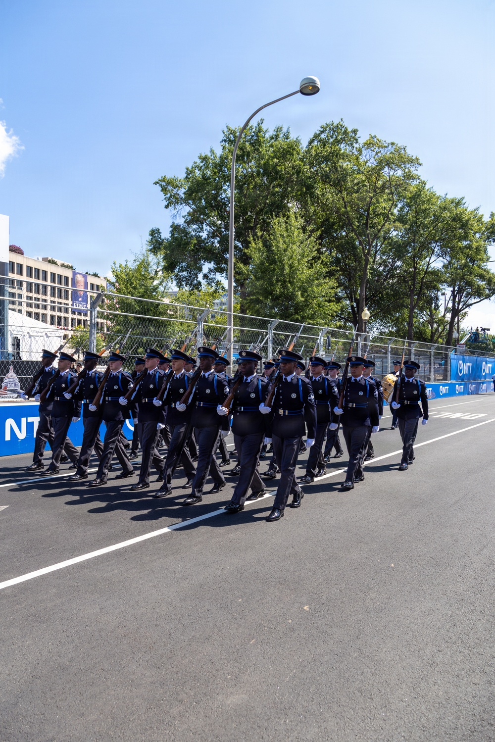 A uniformed marching unit is parading down a closed street lined with blue NTT-branded barriers and chain-link fencing. The personnel are dressed in dark navy uniforms with white gloves and peaked caps, carrying rifles at shoulder arms. The setting appears to be a public event or ceremony in an urban environment, likely Washington D.C., based on visible architecture and signage.