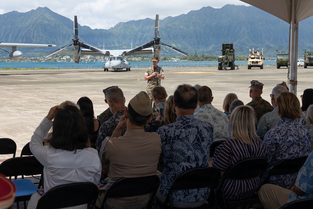 A military briefing or ceremony is taking place on an airfield tarmac, with a uniformed service member addressing a seated audience under a white canopy tent. The backdrop features a V-22 Osprey tiltrotor aircraft, military ground vehicles, a body of water, and dramatic green mountain terrain consistent with Hawaii. The audience consists of a mix of military personnel in uniform and civilians in casual attire.