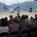 A military briefing or ceremony is taking place on an airfield tarmac, with a uniformed service member addressing a seated audience under a white canopy tent. The backdrop features a V-22 Osprey tiltrotor aircraft, military ground vehicles, a body of water, and dramatic green mountain terrain consistent with Hawaii. The audience consists of a mix of military personnel in uniform and civilians in casual attire.