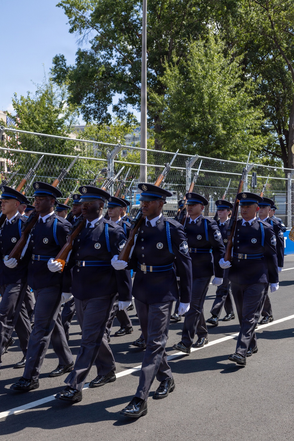 A uniformed honor guard or ceremonial unit is marching in formation during what appears to be a formal parade or official ceremony. The personnel are dressed in matching dark navy blue uniforms with blue accents, white gloves, and peaked caps, carrying rifles at a uniform angle. The setting appears to be an urban street with security fencing and trees visible in the background.