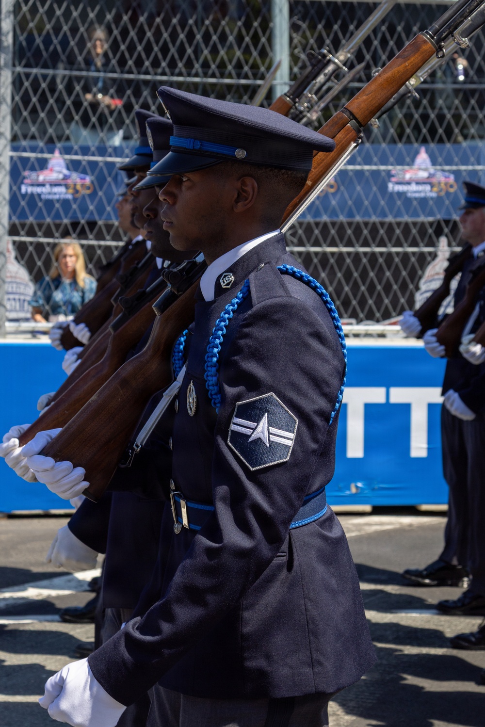 A United States Air Force Honor Guard unit is marching in a formal parade or ceremonial event. The lead airman displays a rank insignia consistent with Airman First Class (A1C) on the sleeve of a dress blue uniform, with blue aiguillette cords and white gloves. A banner in the background reads 'Freedom 250,' suggesting a patriotic or national celebration event.