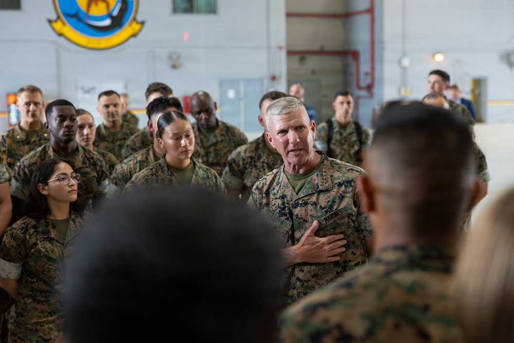 A senior U.S. Marine Corps officer, identifiable by rank insignia and 'MARINES' name tape, addresses a large formation of Marines inside what appears to be a military hangar or bay. The officer has his hand placed over his chest in an emphatic gesture while speaking to the assembled personnel. A military unit insignia/patch is visible on the wall in the background.
