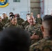 A senior U.S. Marine Corps officer, identifiable by rank insignia and 'MARINES' name tape, addresses a large formation of Marines inside what appears to be a military hangar or bay. The officer has his hand placed over his chest in an emphatic gesture while speaking to the assembled personnel. A military unit insignia/patch is visible on the wall in the background.
