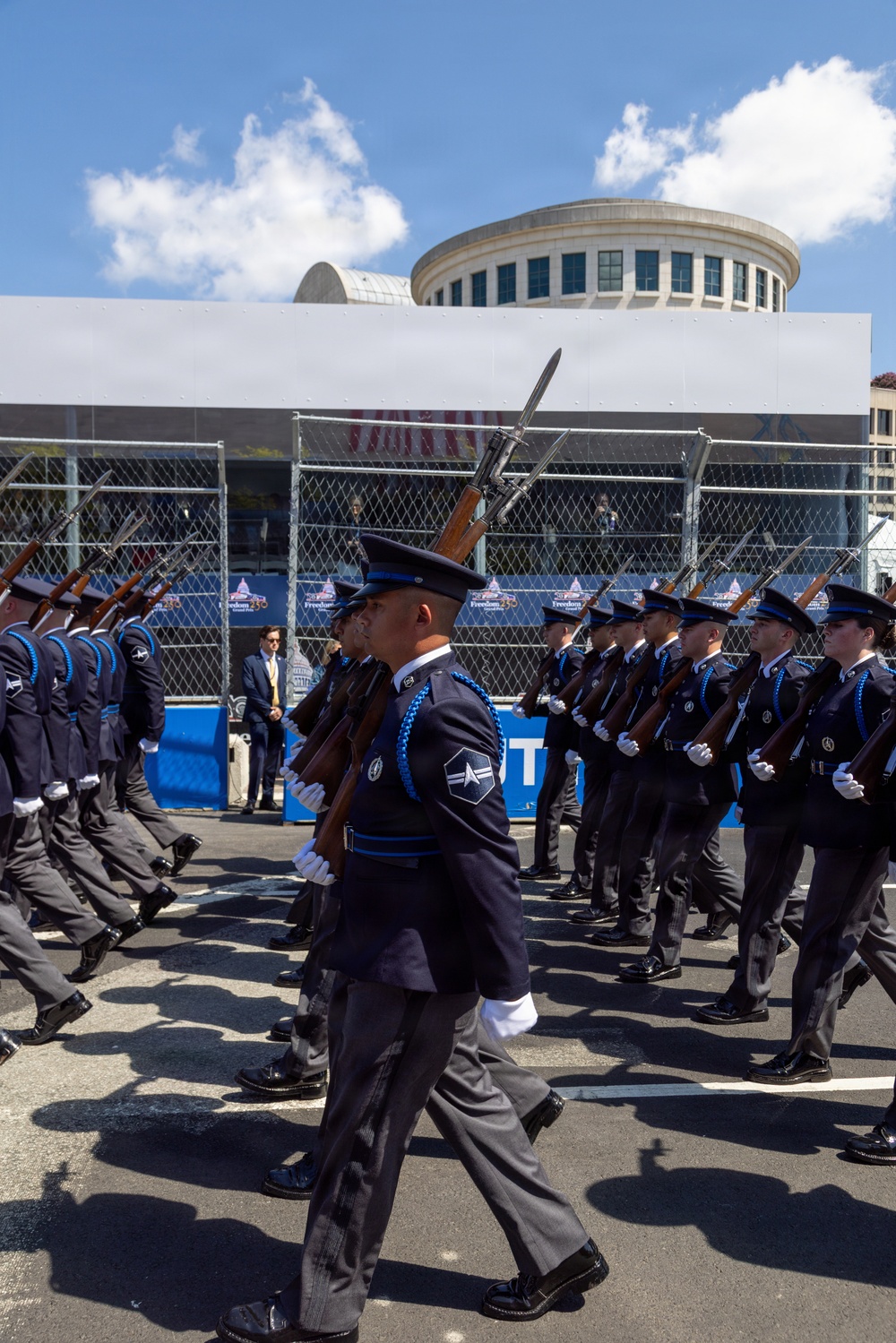 A military honor guard unit is marching in a formal parade, wearing dark dress uniforms with blue aiguillettes and white gloves while carrying rifles with fixed bayonets. The event appears to be a ceremonial parade in an urban setting, with security fencing, signage reading 'Freedom' visible in the background, suggesting a patriotic or national celebration. A circular-topped government or civic building is visible in the background under a partly cloudy sky.