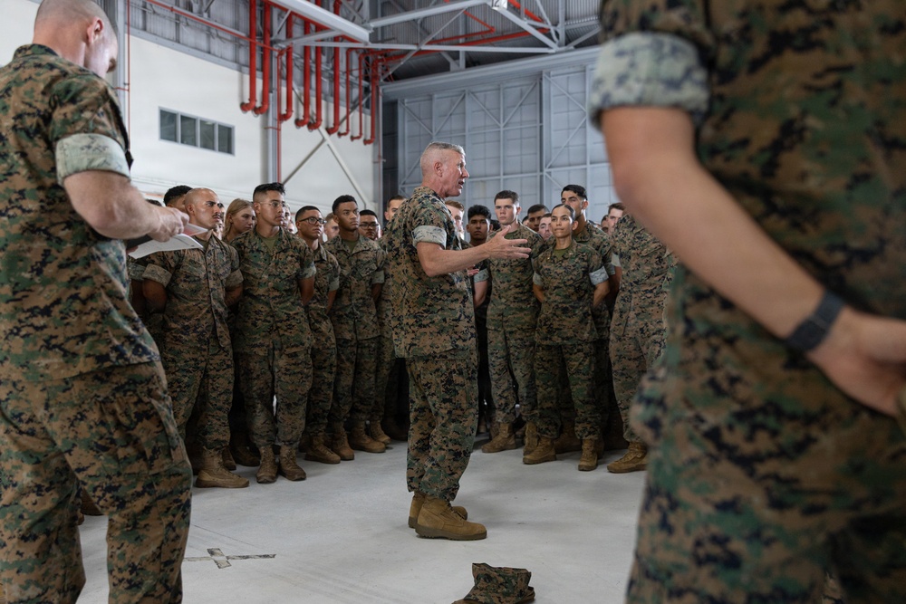 A senior military officer in U.S. Marine Corps MARPAT camouflage uniform addresses a large formation of Marines inside what appears to be a military hangar or large indoor facility. The speaker is positioned centrally, gesturing with his hands while speaking to the assembled group. The Marines are standing at ease in a semicircular formation, attentively listening.