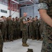 A senior military officer in U.S. Marine Corps MARPAT camouflage uniform addresses a large formation of Marines inside what appears to be a military hangar or large indoor facility. The speaker is positioned centrally, gesturing with his hands while speaking to the assembled group. The Marines are standing at ease in a semicircular formation, attentively listening.