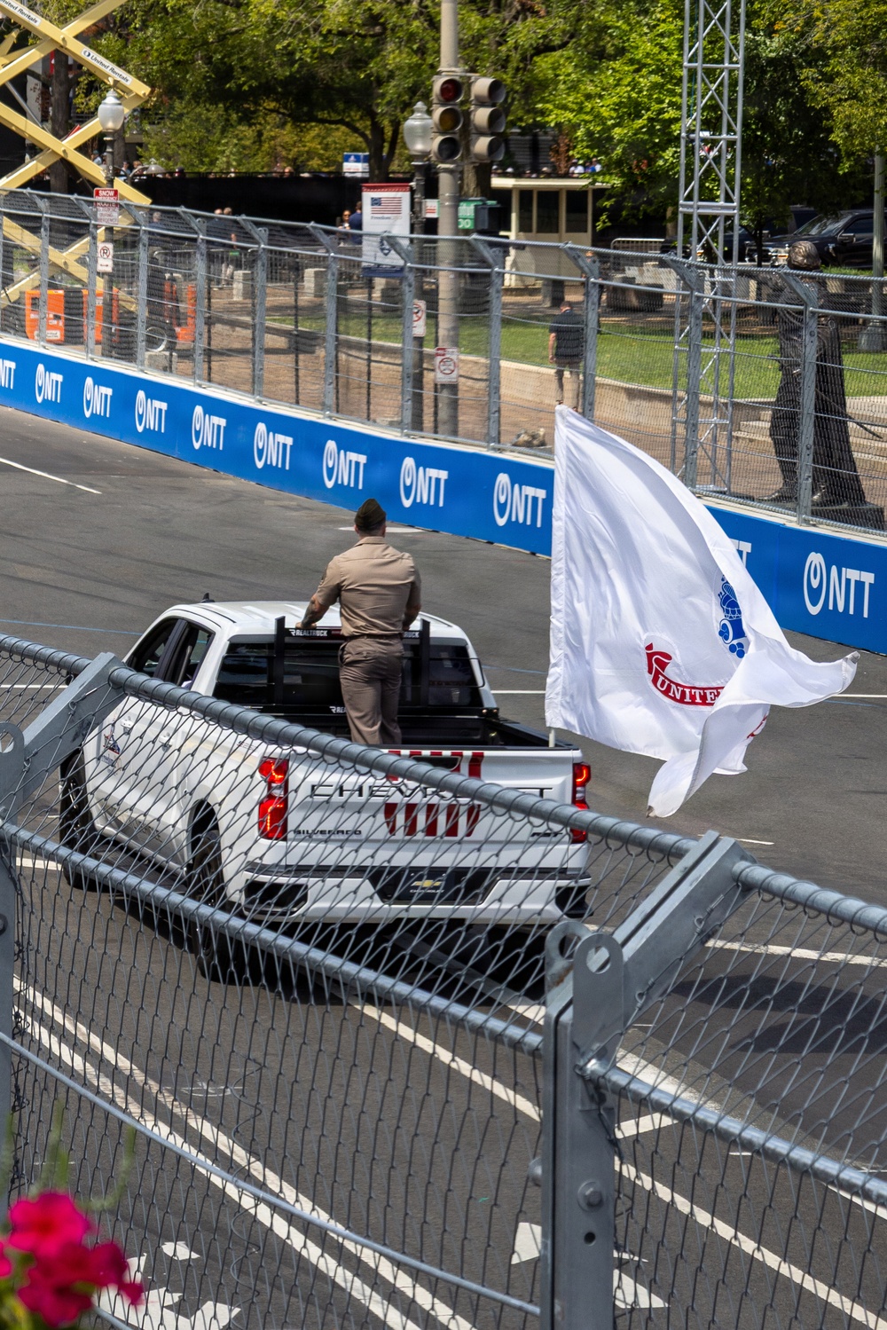 A white Chevrolet Silverado pickup truck is on a temporary street racing circuit, identifiable by NTT IndyCar Series sponsor barriers lining the track. A person in a tan/khaki uniform stands in the truck bed, and a large white flag with partial text reading 'UNITED' is mounted and flying from the vehicle. The setting appears to be an urban street circuit race event, likely the IndyCar Grand Prix of Long Beach or a similar NTT IndyCar Series street race.