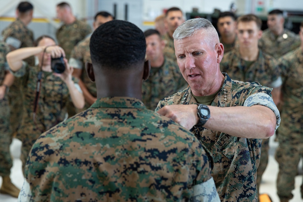 A senior military officer in U.S. Marine Corps MARPAT camouflage uniform is pinning or adjusting something on the collar/shoulder area of another Marine's uniform during what appears to be a formal ceremony. A photographer is capturing the moment in the background, and a large group of Marines in uniform are assembled as witnesses. The setting appears to be an indoor military facility such as a hangar.