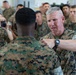 A senior military officer in U.S. Marine Corps MARPAT camouflage uniform is pinning or adjusting something on the collar/shoulder area of another Marine's uniform during what appears to be a formal ceremony. A photographer is capturing the moment in the background, and a large group of Marines in uniform are assembled as witnesses. The setting appears to be an indoor military facility such as a hangar.