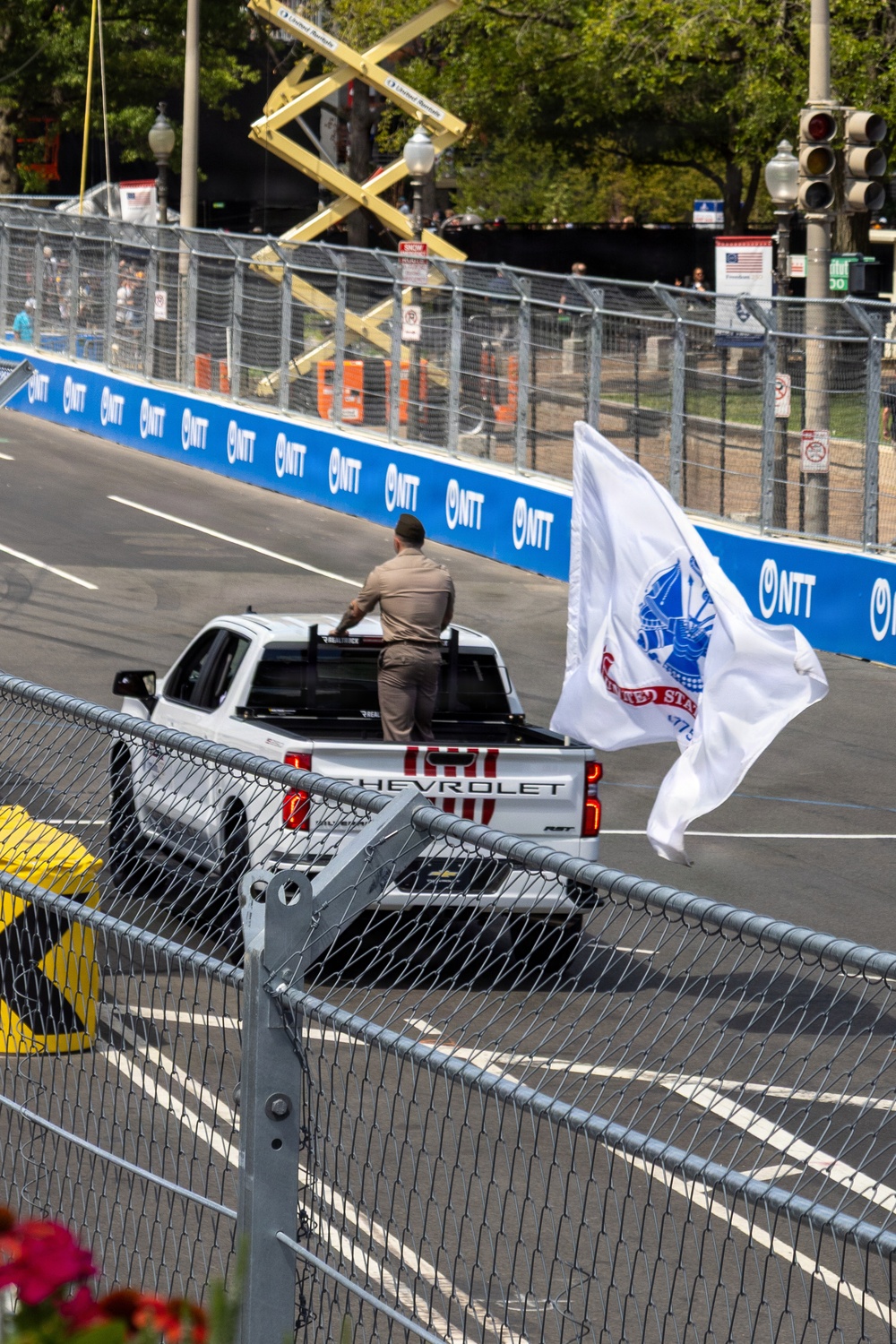 A person in tan/khaki uniform stands in the bed of a white Chevrolet pickup truck on a street racing circuit, holding a large white United States Army flag. The truck is traveling along the track, which is lined with NTT-branded barriers and chain-link fencing. This appears to be a pre-race or ceremonial parade lap at an IndyCar or similar open-wheel racing event.