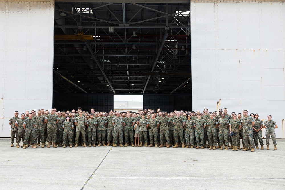 A large group of military personnel, approximately 100-150 individuals, are posed for a formal group photograph in front of a large aircraft hangar. The majority are wearing US Marine Corps MARPAT (woodland digital camouflage) uniforms with tan combat boots. The setting appears to be a military air station or airfield tarmac.