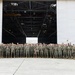 A large group of military personnel, approximately 100-150 individuals, are posed for a formal group photograph in front of a large aircraft hangar. The majority are wearing US Marine Corps MARPAT (woodland digital camouflage) uniforms with tan combat boots. The setting appears to be a military air station or airfield tarmac.