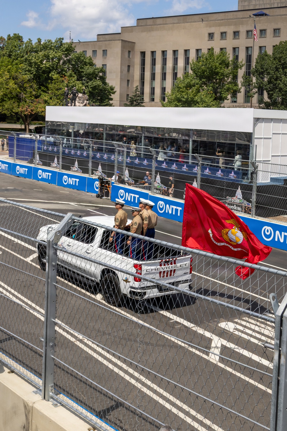 U.S. Marines in dress khaki uniforms ride in the bed of a white Chevrolet pickup truck on a street circuit racing track, carrying the United States Marine Corps flag. The event appears to be an IndyCar or similar motorsport race, evidenced by NTT sponsorship banners lining the barriers. A large neoclassical government building is visible in the background, suggesting a Washington D.C. street circuit venue.