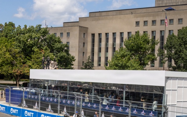 U.S. Marines in dress khaki uniforms ride in the bed of a white Chevrolet pickup truck on a street circuit racing track, carrying the United States Marine Corps flag. The event appears to be an IndyCar or similar motorsport race, evidenced by NTT sponsorship banners lining the barriers. A large neoclassical government building is visible in the background, suggesting a Washington D.C. street circuit venue.