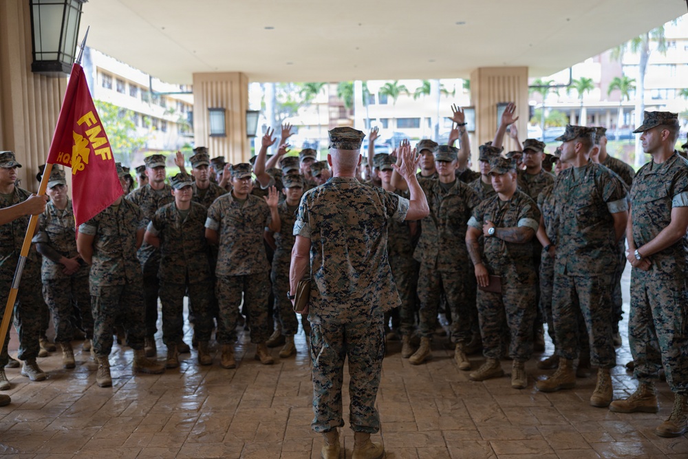 A senior U.S. Marine Corps officer stands with raised hands addressing a large formation of Marines in MARPAT camouflage uniforms under a covered outdoor area. A red FMF HQ guidon flag is visible on the left side of the image. The tropical vegetation and architecture in the background suggest this is likely located at a Hawaii-based installation.