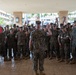 A senior U.S. Marine Corps officer stands with raised hands addressing a large formation of Marines in MARPAT camouflage uniforms under a covered outdoor area. A red FMF HQ guidon flag is visible on the left side of the image. The tropical vegetation and architecture in the background suggest this is likely located at a Hawaii-based installation.