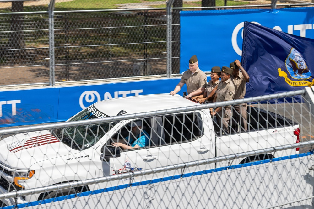 Military personnel in tan/khaki uniforms are riding in the bed of a white Chevrolet Silverado pickup truck at what appears to be a racing event. One person is holding a dark blue United States Navy flag. The truck is traveling along a track area bordered by chain-link fencing and blue NTT sponsor banners.