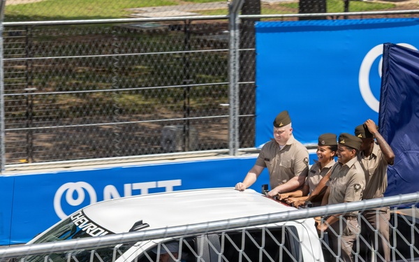 Military personnel in tan/khaki uniforms are riding in the bed of a white Chevrolet Silverado pickup truck at what appears to be a racing event. One person is holding a dark blue United States Navy flag. The truck is traveling along a track area bordered by chain-link fencing and blue NTT sponsor banners.
