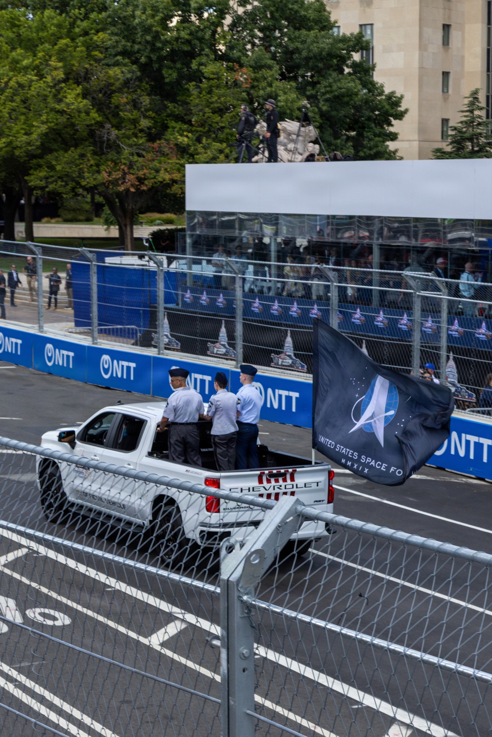 A white Chevrolet Silverado official vehicle carries uniformed United States Space Force personnel along a racing circuit track, flying the USSF flag. The event appears to be an IndyCar or similar open-wheel racing event, evidenced by NTT sponsorship banners lining the track barriers. The setting appears to be an urban street circuit, likely in Washington D.C., based on the neoclassical architecture visible in the background.