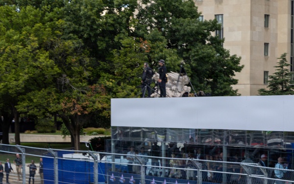 A white Chevrolet Silverado official vehicle carries uniformed United States Space Force personnel along a racing circuit track, flying the USSF flag. The event appears to be an IndyCar or similar open-wheel racing event, evidenced by NTT sponsorship banners lining the track barriers. The setting appears to be an urban street circuit, likely in Washington D.C., based on the neoclassical architecture visible in the background.