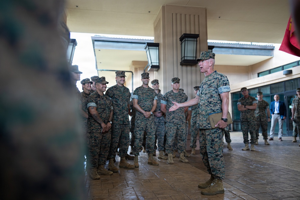 A senior military officer in U.S. Marine Corps digital camouflage (MARPAT) uniform is addressing a group of assembled Marines outdoors under a covered walkway. The speaker appears to be gesturing while speaking, with the assembled Marines listening attentively, some visibly smiling. The setting appears to be a military installation building with a tan/beige exterior.