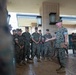 A senior military officer in U.S. Marine Corps digital camouflage (MARPAT) uniform is addressing a group of assembled Marines outdoors under a covered walkway. The speaker appears to be gesturing while speaking, with the assembled Marines listening attentively, some visibly smiling. The setting appears to be a military installation building with a tan/beige exterior.