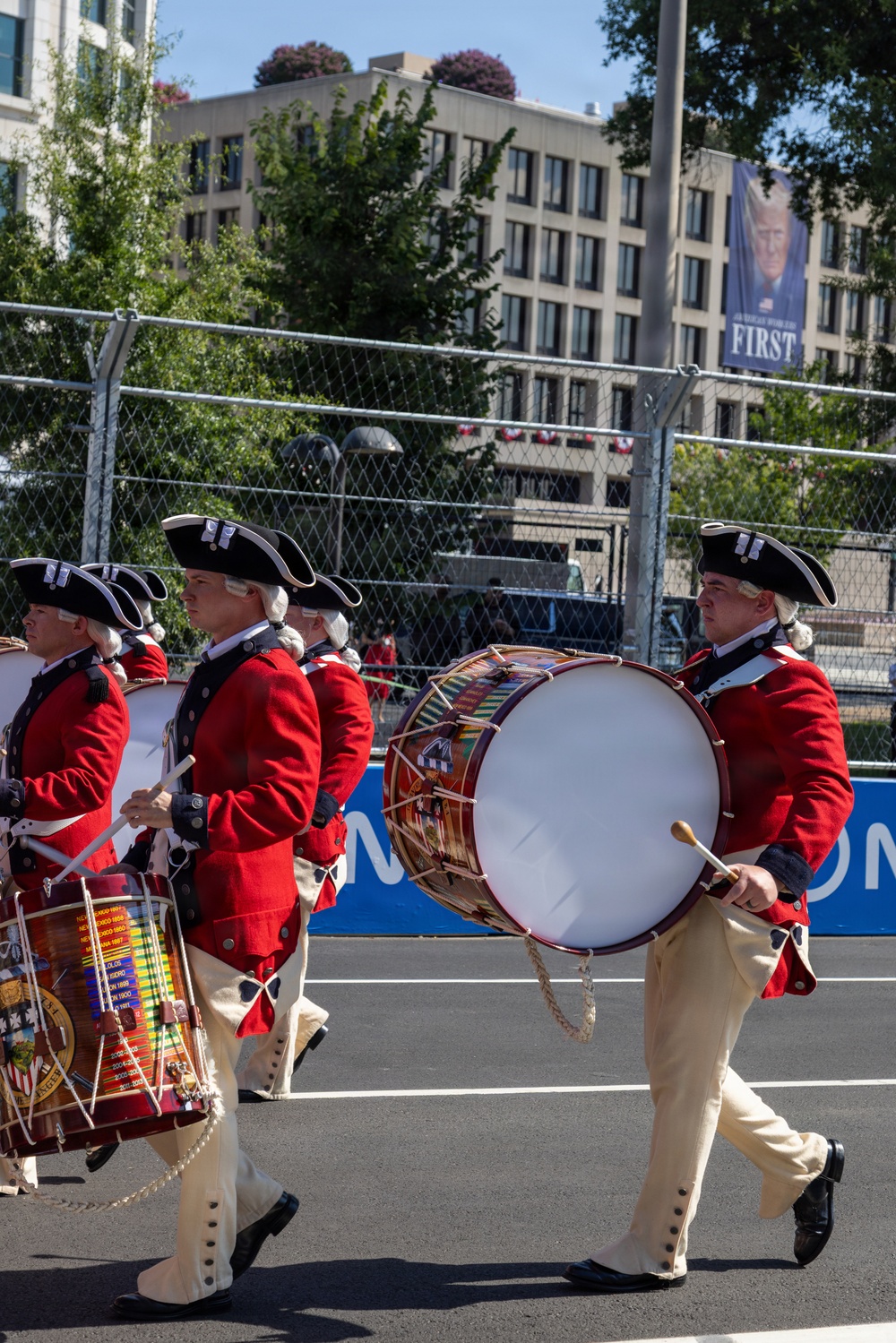A colonial-era style military drum corps marches in formation along a paved route, wearing 18th-century Revolutionary War period uniforms consisting of red coats, white breeches, and tricorn hats. The drummers carry ornately decorated snare and bass drums while marching past a chain-link security fence. In the background, a banner on a building reads 'FIRST,' suggesting a political or ceremonial event context.