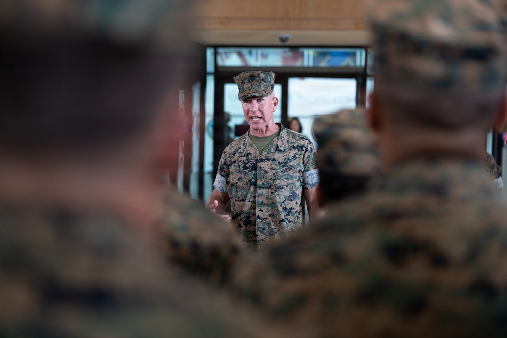 A U.S. Marines officer in MARPAT woodland camouflage uniform is addressing a group of Marines in what appears to be an indoor facility. The central figure appears to be a senior officer speaking to assembled personnel, with other uniformed Marines visible in the foreground and background. The image uses a shallow depth of field, keeping the speaking officer in focus while blurring surrounding figures.