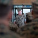 A U.S. Marines officer in MARPAT woodland camouflage uniform is addressing a group of Marines in what appears to be an indoor facility. The central figure appears to be a senior officer speaking to assembled personnel, with other uniformed Marines visible in the foreground and background. The image uses a shallow depth of field, keeping the speaking officer in focus while blurring surrounding figures.