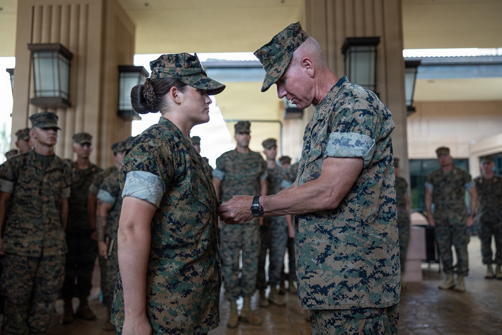 A military award or decoration ceremony is taking place, with a senior male officer pinning a medal or insignia onto a female service member's uniform. Both individuals are wearing US Marine Corps MARPAT (digital camouflage) uniforms. A formation of Marines stands at attention in the background, observing the ceremony.