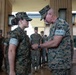 A military award or decoration ceremony is taking place, with a senior male officer pinning a medal or insignia onto a female service member's uniform. Both individuals are wearing US Marine Corps MARPAT (digital camouflage) uniforms. A formation of Marines stands at attention in the background, observing the ceremony.
