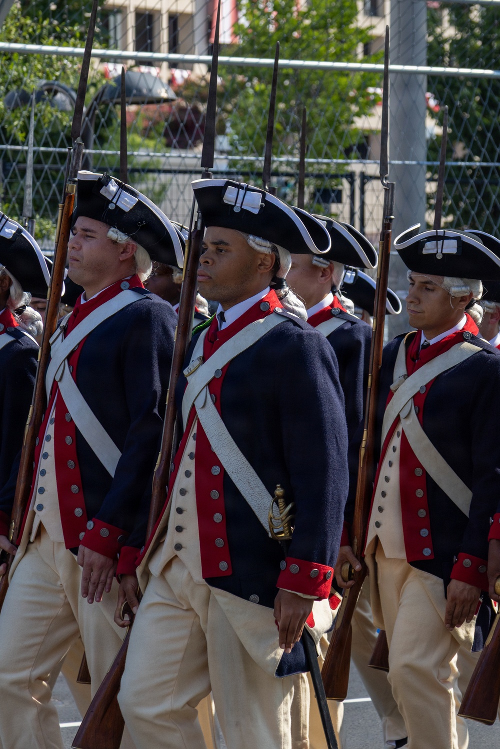 A group of soldiers in American Revolutionary War-era uniforms are marching in formation during what appears to be a ceremonial parade or reenactment. They wear matching dark blue coats with red facings, white crossbelts, buff/cream breeches, and tricorn hats with white trim. Each soldier carries a musket with a fixed bayonet at shoulder arms.