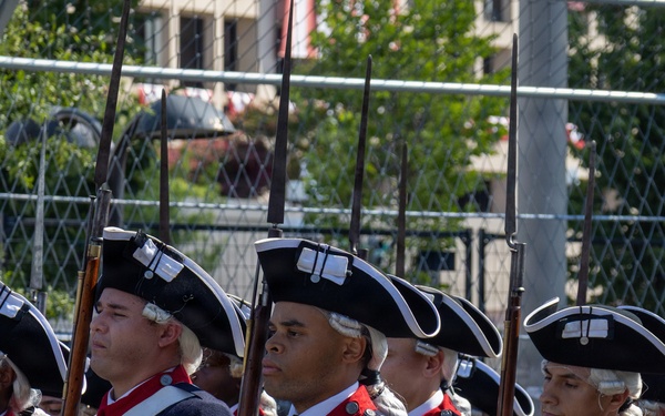 A group of soldiers in American Revolutionary War-era uniforms are marching in formation during what appears to be a ceremonial parade or reenactment. They wear matching dark blue coats with red facings, white crossbelts, buff/cream breeches, and tricorn hats with white trim. Each soldier carries a musket with a fixed bayonet at shoulder arms.