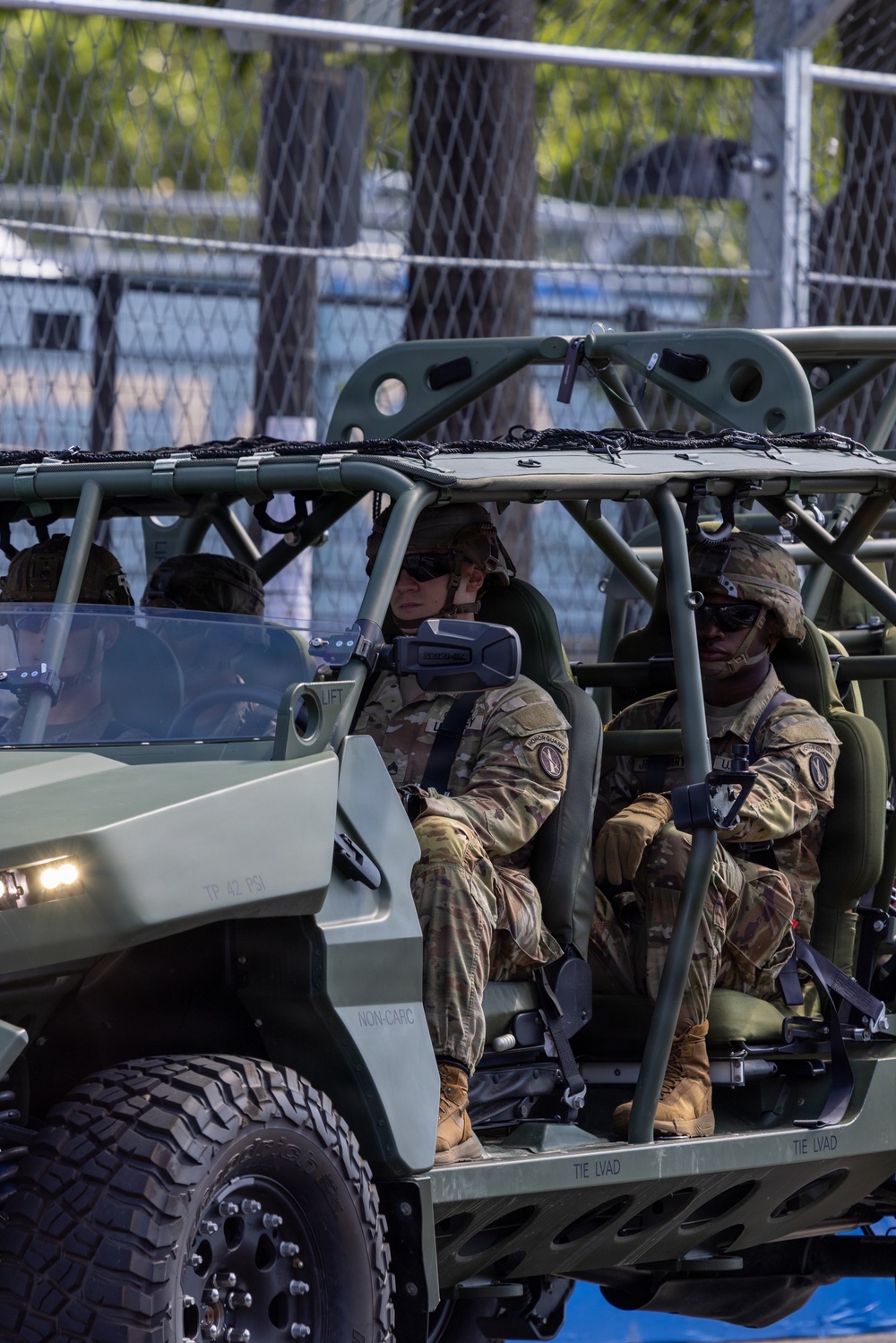 The image shows U.S. Army soldiers seated in a light tactical vehicle, identifiable as an Infantry Squad Vehicle (ISV) based on its olive-drab open-frame roll cage design. The soldiers are wearing MultiCam (OCP) uniforms, combat helmets, sunglasses, and tactical gloves. The vehicle displays markings including 'NON-CARC', 'TIE LVAD', and 'TP 42 PSI', consistent with U.S. Army vehicle labeling standards.