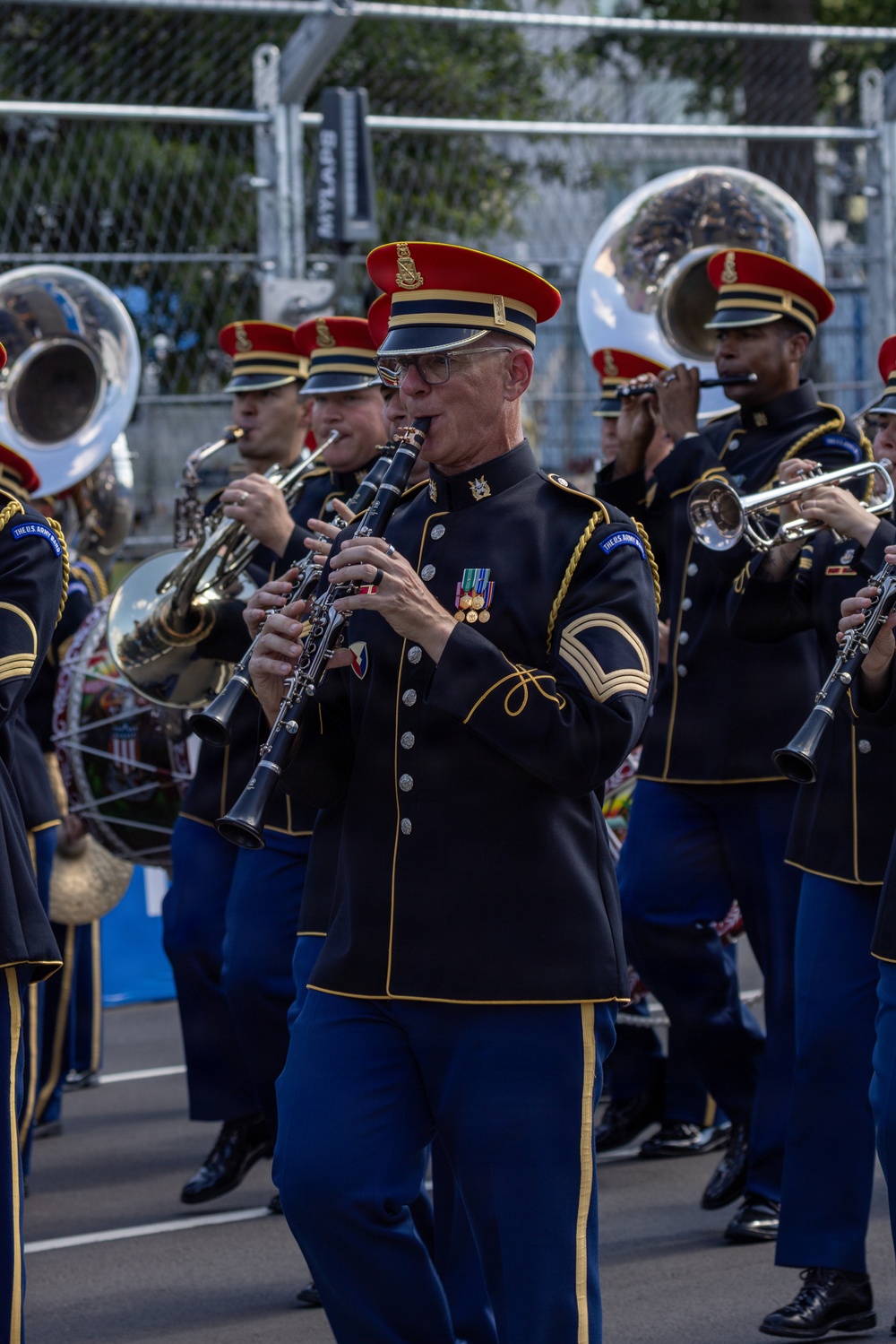 A U.S. Army marching band performs in what appears to be a parade or public event. The musicians are dressed in formal ceremonial uniforms featuring dark navy jackets with gold trim, blue trousers, and distinctive red-topped caps. The foreground features a clarinetist with visible rank insignia and service medals, surrounded by bandmates playing brass instruments including tubas and trumpets.